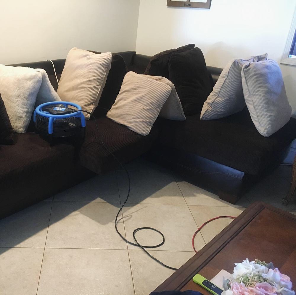 A blue air mover running on a tile floor beside a sofa, drying the room after cleaning.